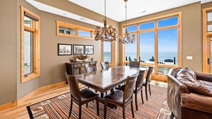 A dining area with a large wooden table, chairs, and a chandelier. There are large windows with a view of the ocean. - Lakeview 5 bedroom Executive Suite (Two Harbors)