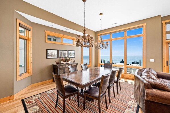 A dining area with a large wooden table, chairs, and a chandelier. There are large windows with a view of the ocean.