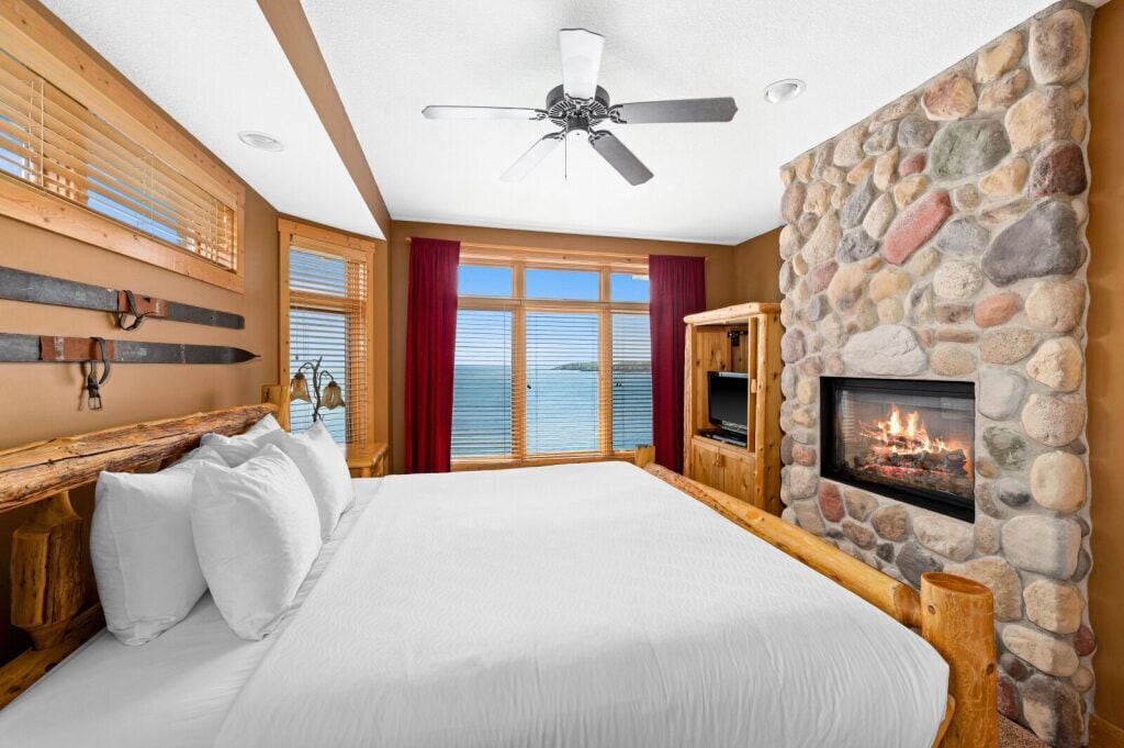 Ein Schlafzimmer mit einem großen Bett, einem Kamin aus Stein, einem Deckenventilator und Meerblick durch große Fenster.