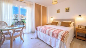 Een hotelkamer met een bed, een eettafel met stoelen, een balkon met gordijnen en uitzicht op gebouwen.