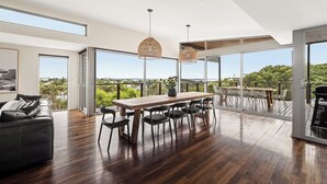 Une pièce spacieuse avec une table et des chaises de salle à manger en bois, un canapé noir et de grandes fenêtres donnant sur un paysage verdoyant et des bâtiments.