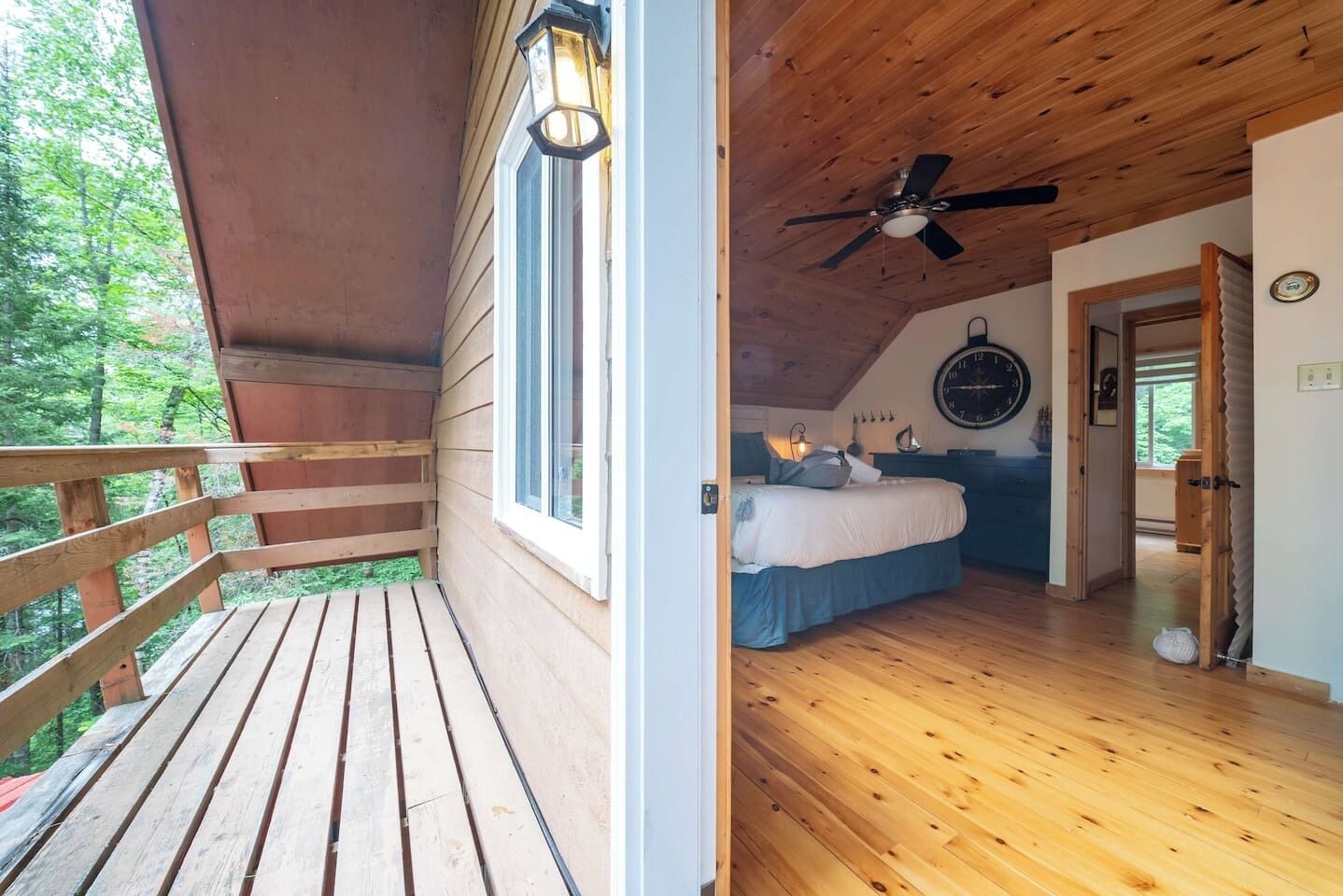 A wooden deck with a railing, a bedroom with a bed, a ceiling fan, and a clock on the wall.