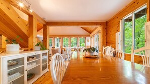 A wooden interior with a long dining table, white chairs, and large windows offering a view of greenery. - Lakeside Retreat  with Hot Tub and Bunkie. Sleeps 10 (Otter Lake)
