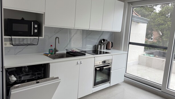 A modern kitchen with white cabinetry, a Bosch dishwasher, a built-in oven, and a large window offering a view of the outdoors.