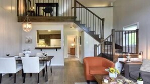 A spacious living area with a dining table, orange sofa, and a wooden coffee table with snacks and drinks. A staircase connects to an upper level with a balcony.