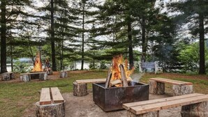 Een camping met meerdere vuurplaatsen, omringd door houten banken en picknicktafels, gelegen in een bebost gebied met bomen en een meer op de achtergrond.