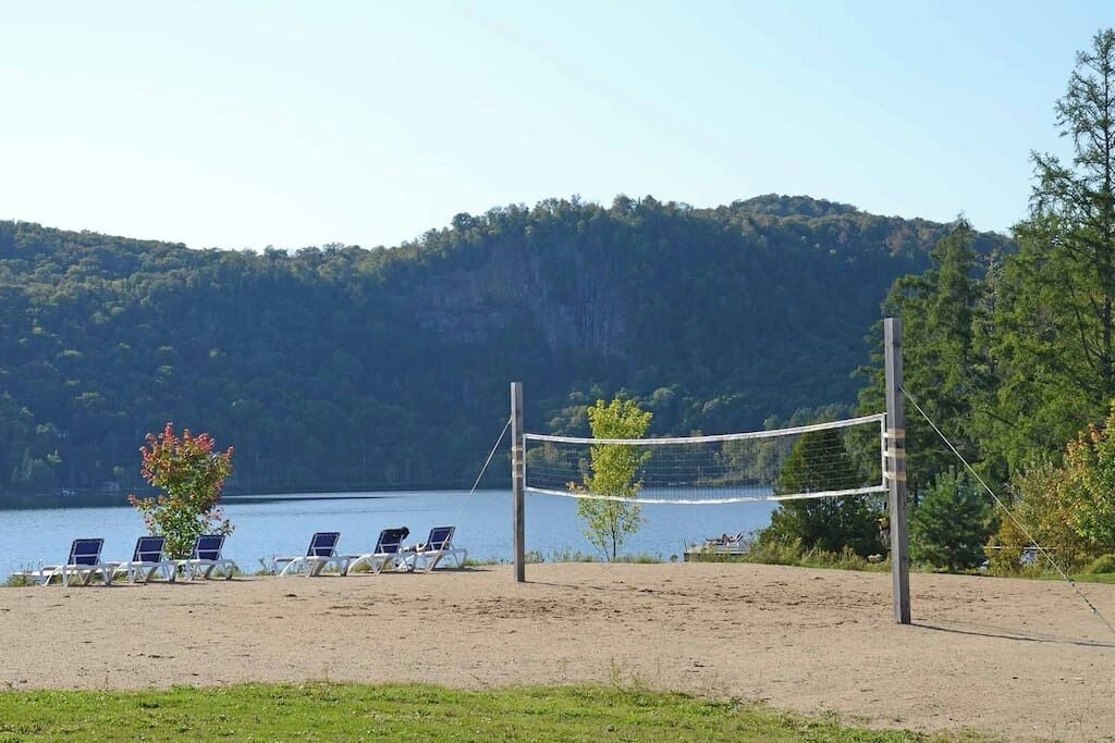 Een strandvolleybalveld met stoelen en een net, uitkijkend over een meer met een bebost heuvel op de achtergrond.