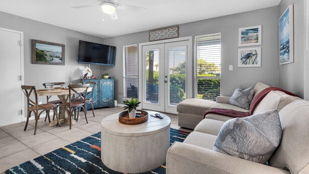 A living room with a sofa, coffee table, and TV. There is a dining area with chairs and a table. The room has a ceiling fan and a view through the glass doors. - Cozy 1-bedroom condo in wonderful Destin with AC (Destin)