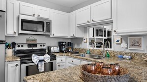 A kitchen with white cabinets, a microwave, stove, and a granite countertop. There are starfish decorations on the cabinets and a sign that says "Beach Rules". - Cozy 1-bedroom condo in wonderful Destin with AC (Destin)