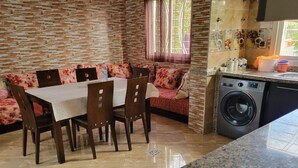 Un coin repas avec une table et des chaises, un canapé agrémenté de coussins à motifs floraux, un lave-linge et un plan de travail de cuisine.