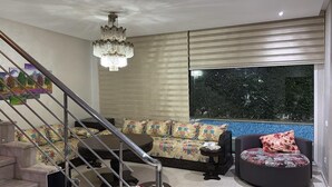 Un salon moderne avec un escalier, un lustre, un canapé agrémenté de coussins à motifs floraux, une table basse ronde et une vue sur une piscine aux carreaux bleus.