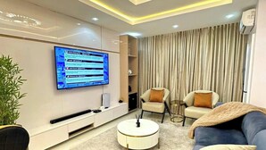 A modern living room with a flat-screen TV mounted on the wall, a round coffee table, a blue sofa, and two armchairs. - 4th Manor Shortlets (Lekki)