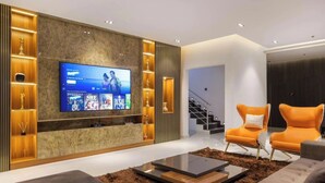 A modern living room with a large flat-screen TV, two orange armchairs, a glass coffee table, and a staircase in the background. - 4th Manor Shortlets (Lekki)