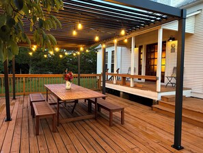 Una terraza de madera con una mesa, bancos, luces colgantes y una zona cubierta con barandilla.
