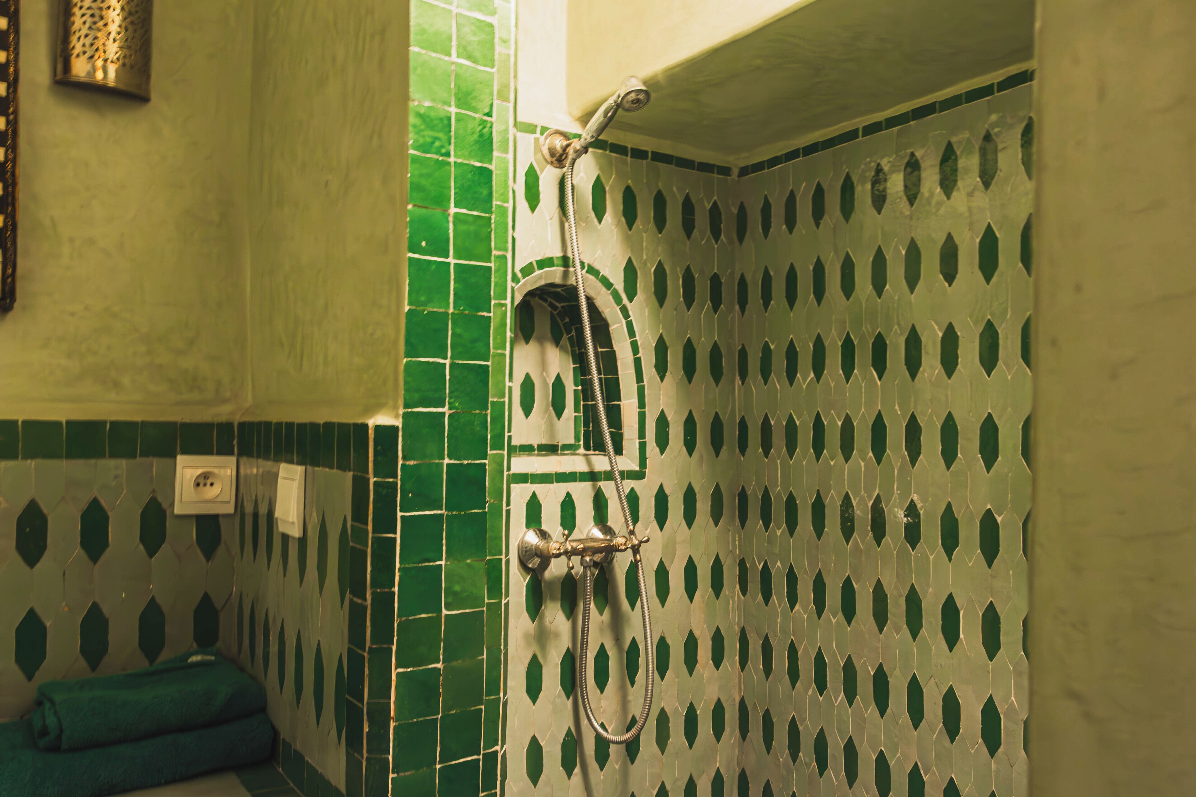 A bathroom with green and white patterned tiles, a shower with a handheld showerhead, and a towel rack with folded towels.