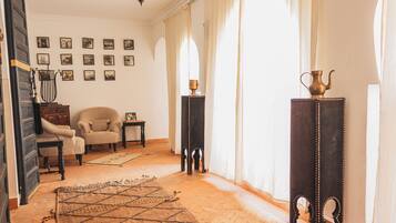 A room with a patterned rug, two armchairs, a small table with a lamp, a tall stand with a vase, and a wall with framed pictures.