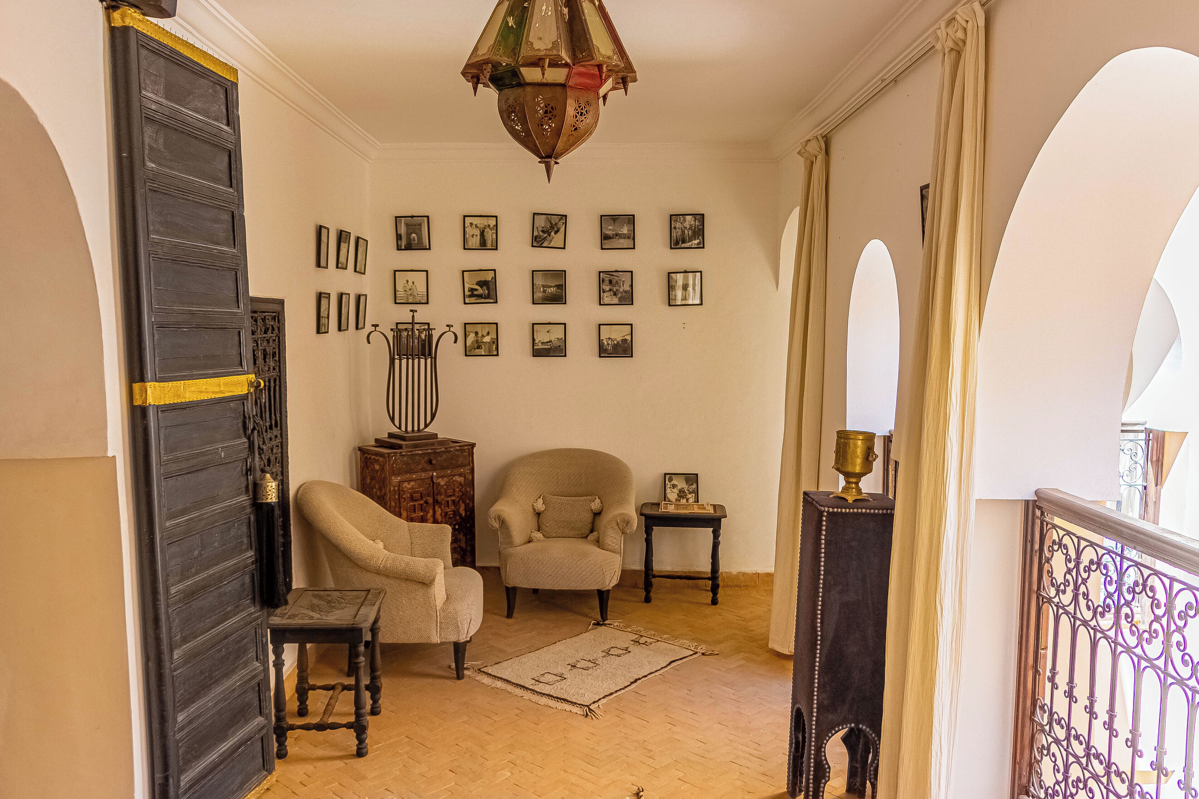 A room with a wooden floor, a large dark wooden door, a beige armchair, a smaller armchair, a small table with a lamp, and a wall with framed pictures.