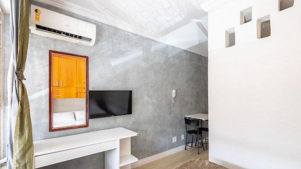 A modern room with a grey textured wall, a mounted flat-screen TV, a wooden wardrobe, and a white desk. - Cozy Studio in Asa Norte BSB Esz0103 (Brasília)