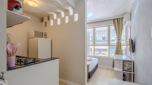 A compact kitchen with a microwave, refrigerator, and gas stove. A bed is visible through a window, and a TV is mounted on the wall. - Cozy Studio in Asa Norte BSB Esz0103 (Brasília)