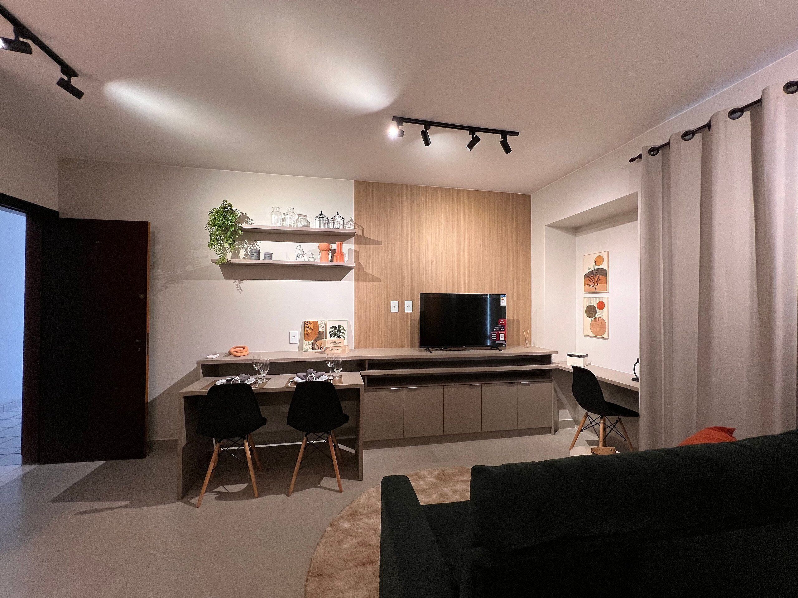 Un salon moderne doté d’un téléviseur à écran plat, d’un coin repas avec des chaises et d’un espace cuisine avec un meuble et des étagères en bois.