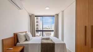 A hotel room with a bed, a nightstand, a remote control, a window with curtains, and a view of high-rise buildings. - Comfort and Convenience in Brasilia - Get0302 (Guará)