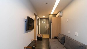 A narrow hallway leading to a room with a bed, a TV mounted on the wall, a wooden bench, and a sofa. - Grand Mercure Brasilia 5 Estrelas - Gmb0414 (Brasília)
