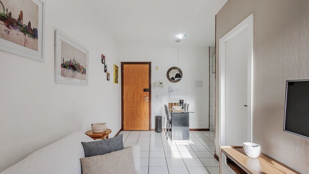 A hallway with a wooden door, a small table with a basket, a sofa with cushions, a coffee machine, and a TV mounted on the wall. - Charming Apartment in Asa Norte, Brasilia Gpk0115 (Brasília)