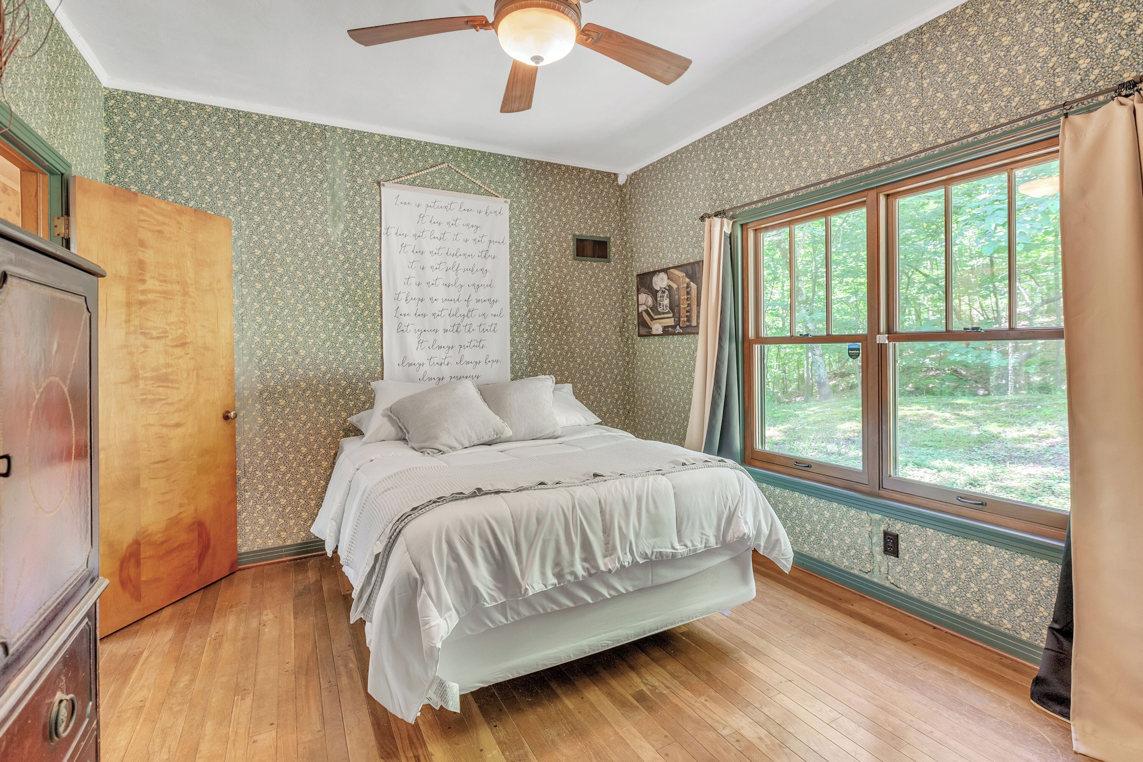 A bedroom with a wooden floor, a bed with white bedding, a ceiling fan, a window with curtains, and a wall with a floral pattern.