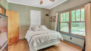 A bedroom with a wooden floor, a bed with white bedding, a ceiling fan, a window with curtains, and a wall with a floral pattern.