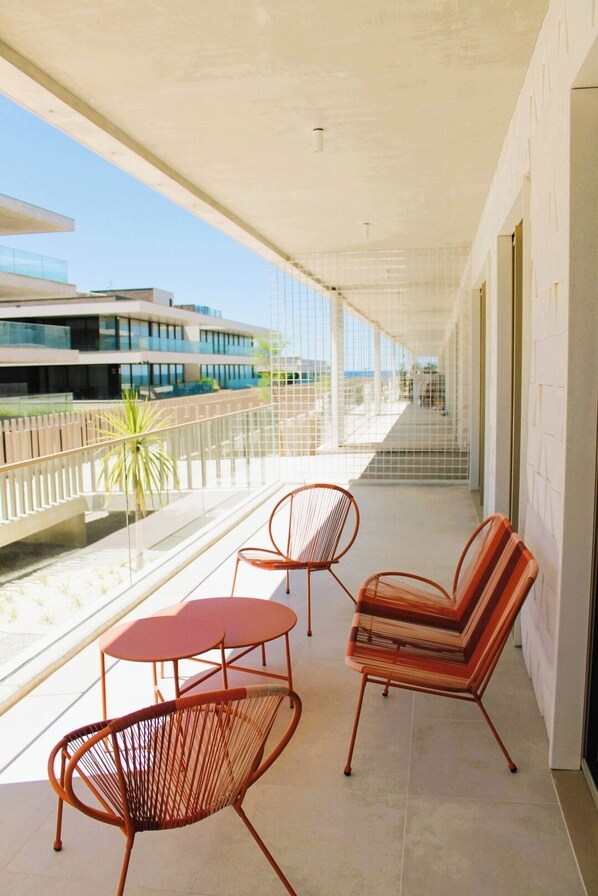 Varanda com duas cadeiras vermelhas e uma mesa pequena, com vista para um edifício moderno e um corpo de água.