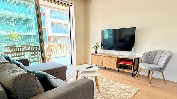 Un salon moderne avec un canapé gris, une petite table blanche et un fauteuil gris. Un téléviseur à écran plat est fixé au mur, au-dessus d’un meuble en bois.