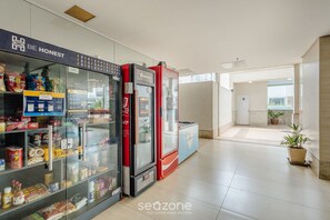 A hallway with vending machines stocked with snacks and beverages, a sign that says "BE HONEST", and a potted plant in the distance. - Beautiful Studios in Condominium With Pool - Aguas Claras - Madb (Águas Claras)