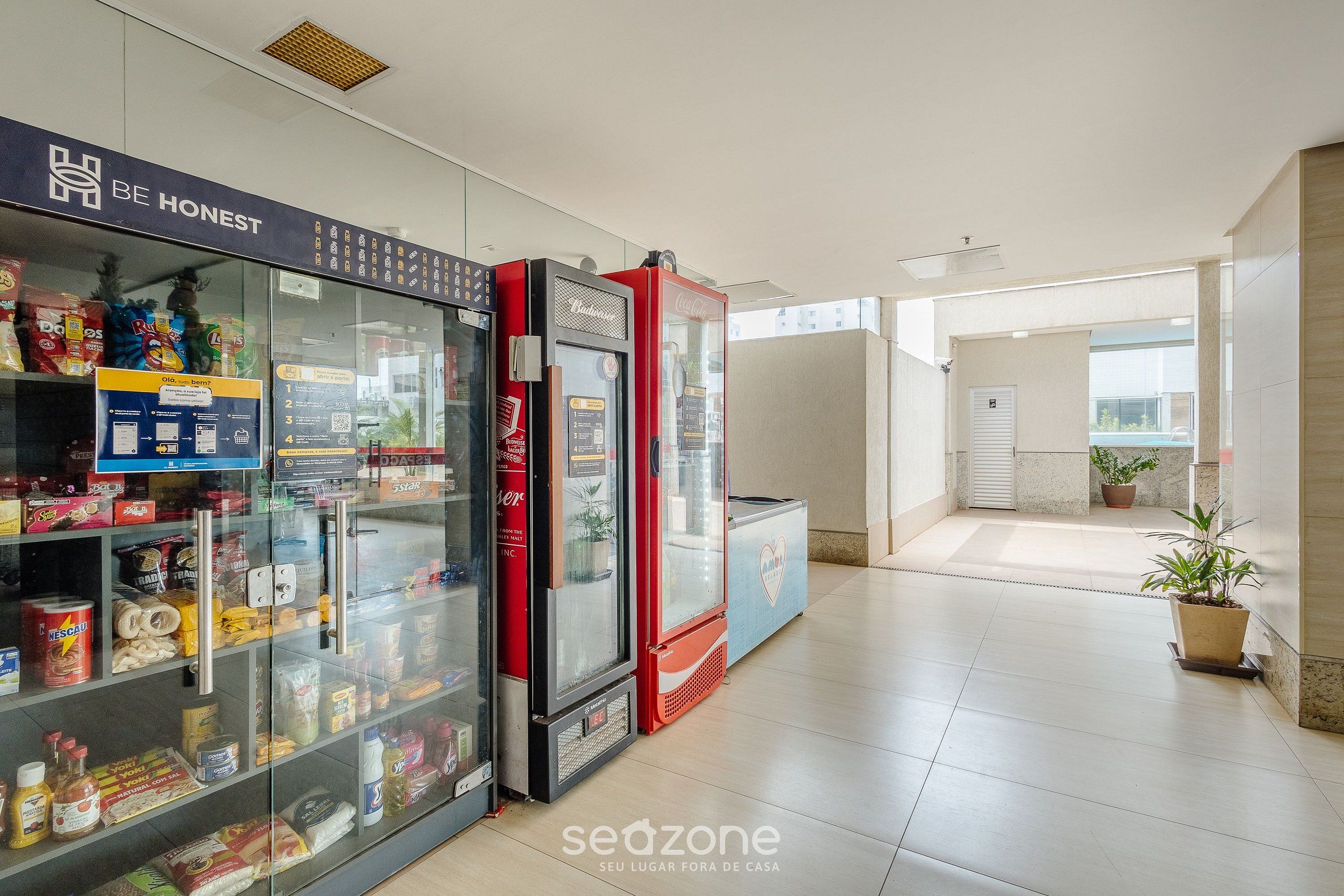 A hallway with vending machines stocked with snacks and beverages, a sign that says "BE HONEST", and a potted plant in the distance.