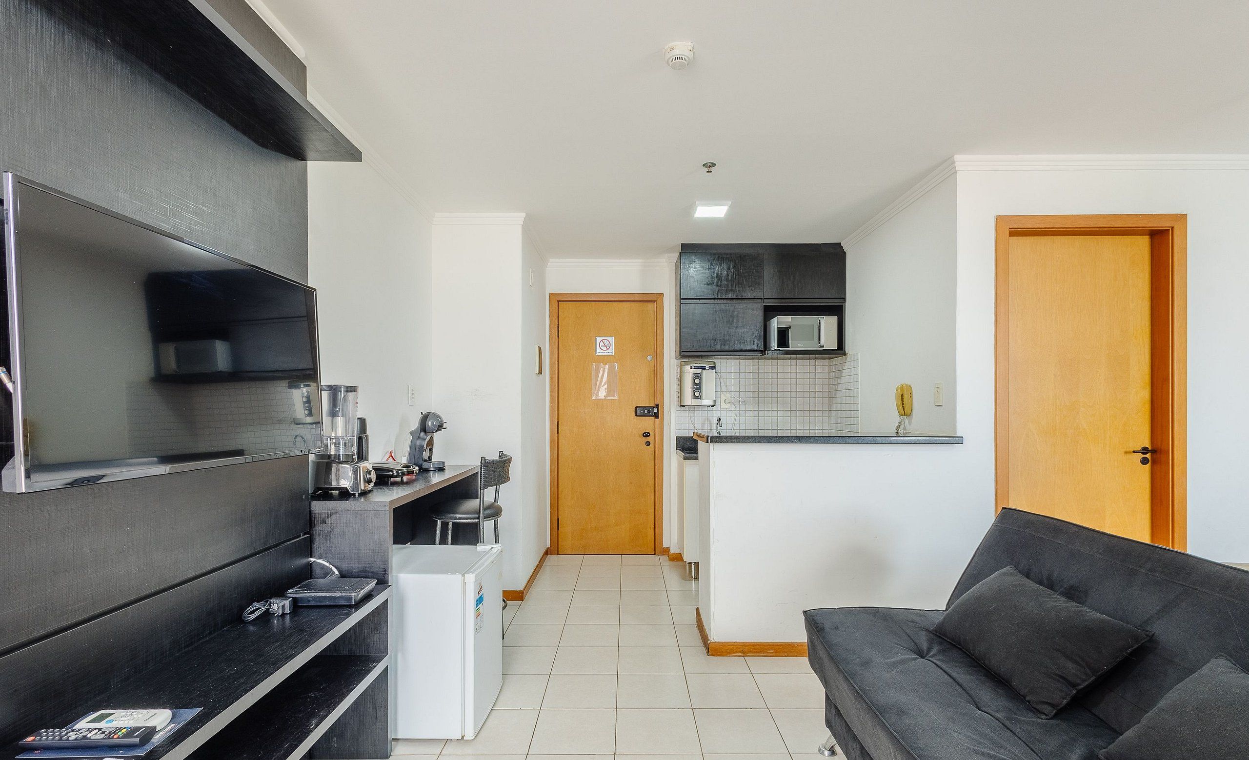 A modern kitchen with a microwave, toaster, and coffee maker. There is a black sofa and a wooden door in the living area.