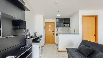 A modern kitchen with a microwave, toaster, and coffee maker. There is a black sofa and a wooden door in the living area.