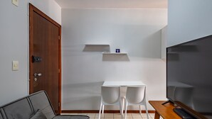 Ein kleiner, moderner Raum mit einer Holztür, einem grauen Sofa, einem weißen Tisch mit zwei Stühlen, einem Fernseher und einem Regal mit einem kleinen Gegenstand darauf.