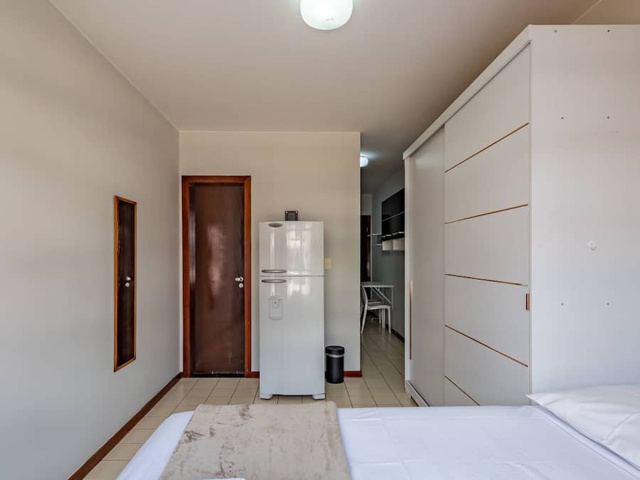 A small, narrow hotel room with a bed, a wardrobe, a door, a refrigerator, and a trash bin.