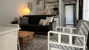 A modern living room with a black sofa, a white armchair, a wooden coffee table, and a patterned rug. There is a kitchen area with shelves and a microwave visible in the background. - A-Chic 1-bedroom apartment with WiFi, AC in vibrant Istanbul (İstanbul)