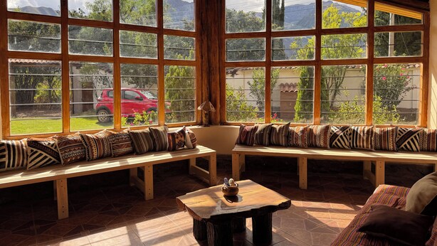 A cozy room with large windows, a wooden bench, a small table, and a view of a red car outside. - Andean Cottage w/ Garden & Workspace near Urubamba (Urubamba)