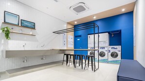 A modern laundry facility with a white tiled wall, a blue accent wall, and a bar area with stools. There are three washing machines and a dryer. - Noas - Apartments in the South Zone of SP (São Paulo)