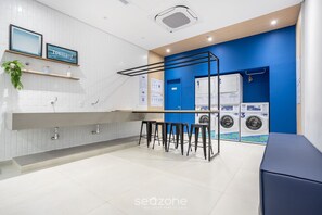 Een moderne wasruimte met een witte betegelde muur, een blauwe accentmuur en een bar met krukken. Drie wasmachines en een droger zijn aanwezig.