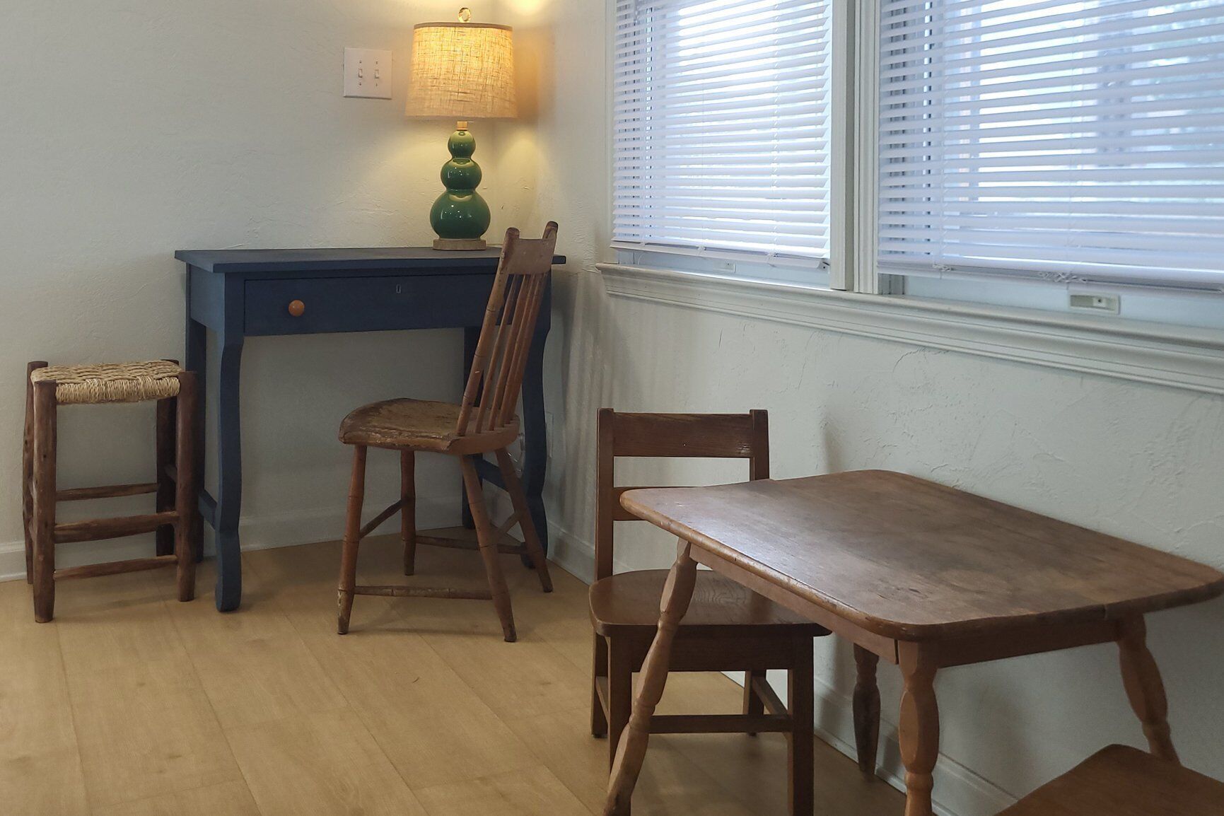A room with a wooden desk, two chairs, a small table, and a lamp.