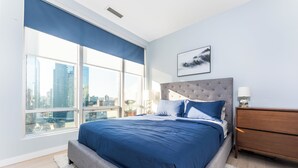 A modern bedroom with a large bed, a grey upholstered headboard, a white dresser, and a city view through floor-to-ceiling windows. - Luxury Executive Downtown 1 bedroom Suite (Vancouver)