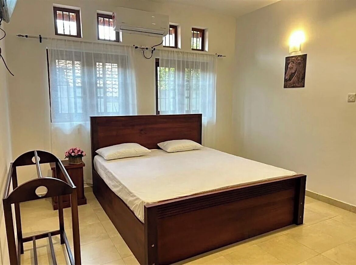 Un dormitorio con una cama de madera, una silla, una mesita con un florero y una lámpara fijada a la pared.
