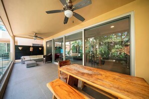 A spacious balcony with a wooden table and benches, a ceiling fan, and a view of the outdoor area.