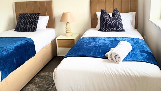 Une chambre d’hôtel avec deux lits, chacun recouvert d’une couvre-lit bleue et doté de draps blancs. Il y a une table de chevet avec une lampe entre les deux lits, et une œuvre d’art est accrochée au mur au-dessus des lits.