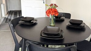 Une table à manger avec des assiettes noires et un vase de fleurs. Des chaises sont disposées autour de la table, et une porte blanche est visible à l’arrière-plan.