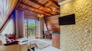 A cozy room with a wooden ceiling, a flat-screen TV mounted on a textured wall, a dining area with a table and chairs, and a large window with curtains. - Fully Equipped Studios Near Armacao Beach and Beto Carrero Park - TRS (Penha)
