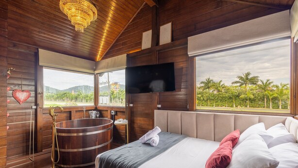 A wooden room with a bed, a TV, a large window, and a bathtub. - Fully Equipped Studios Near Armacao Beach and Beto Carrero Park - TRS (Penha)