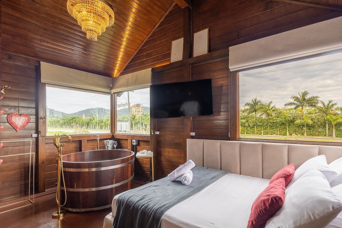 A wooden room with a bed, a TV, a large window, and a bathtub.